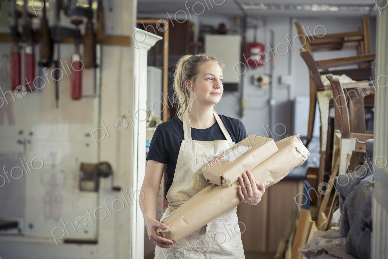 A young woman in a workshop, holding two packages wrapped in brown packaging paper,…