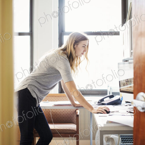 A young woman in an office standing over a desk and working at a computer.