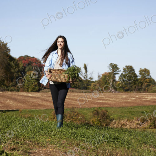 A young woman in working clothes walking across a field, holding a basket of crops. A young woman in working clothes walking across a field, holding a basket of crops.