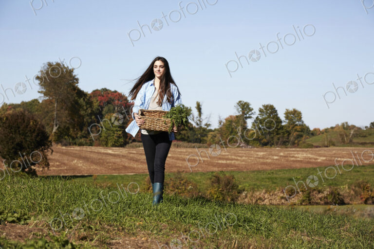 A young woman in working clothes walking across a field, holding a basket of crops.