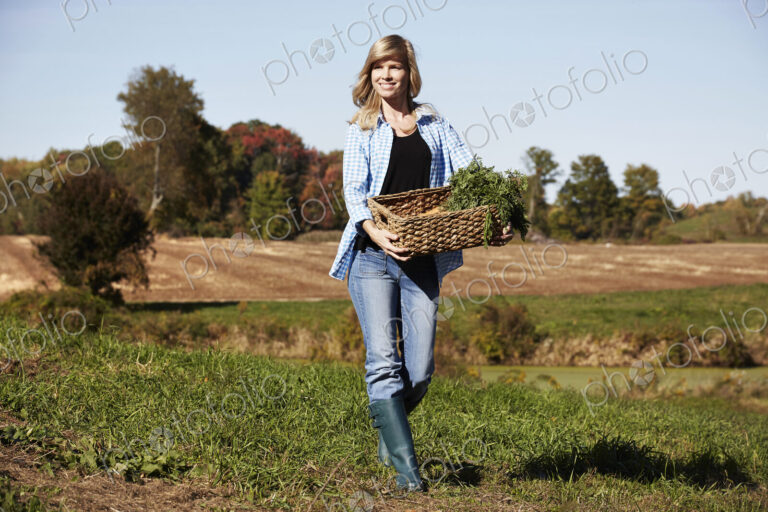 A young woman in working clothes walking across a field, holding a basket of crops.