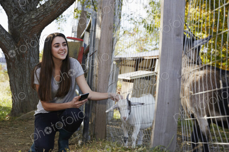 A young woman kneeling down and petting a kid goat through a wire fence.