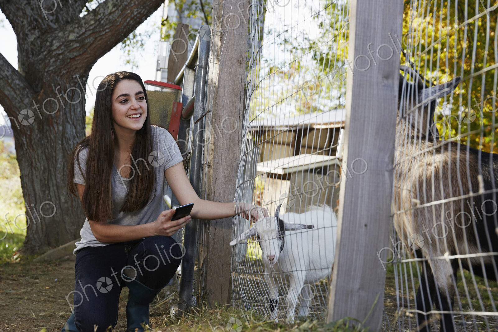 A young woman kneeling down and petting a kid goat through a wire fence.