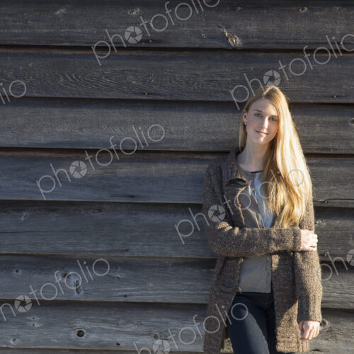 A young woman leaning against the wooden wall of a barn wearing a long knitted coat.