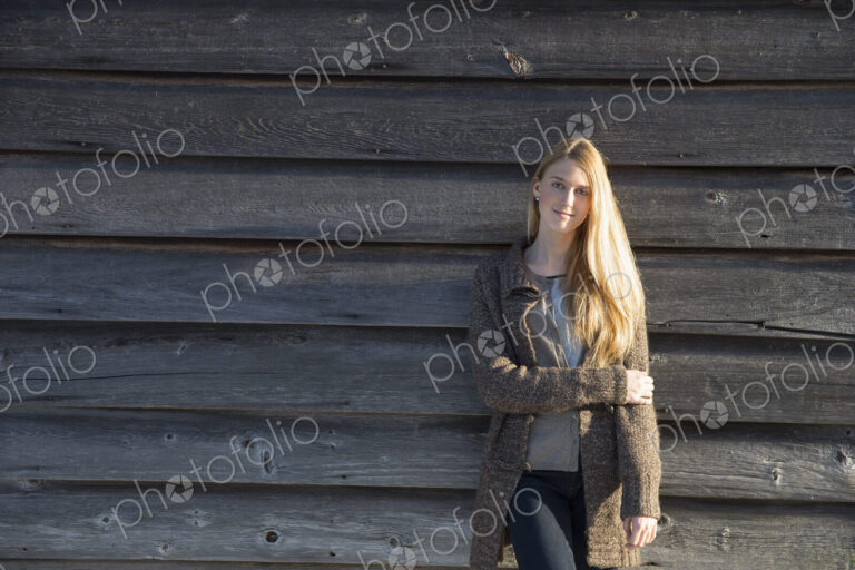 A young woman leaning against the wooden wall of a barn wearing a long knitted coat.