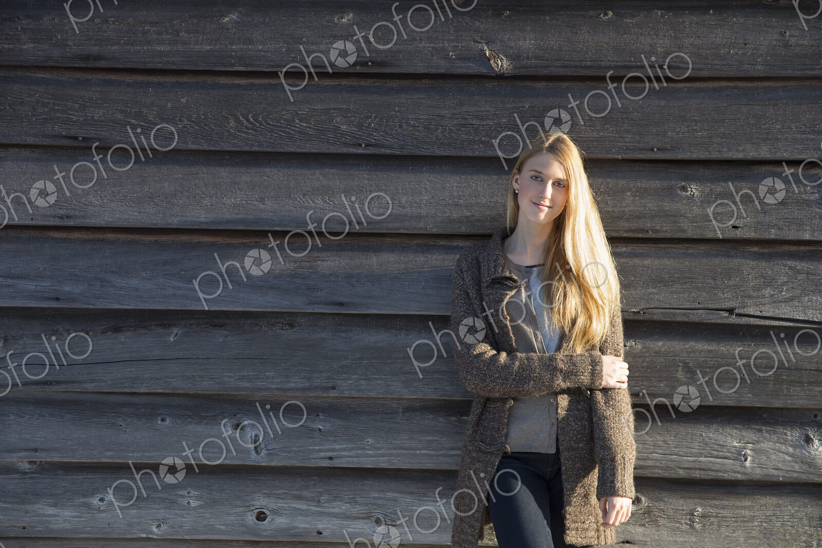 A young woman leaning against the wooden wall of a barn wearing a long knitted coat.