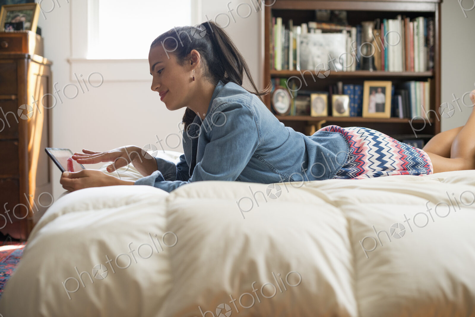 A young woman lying on her bed using a digital tablet.