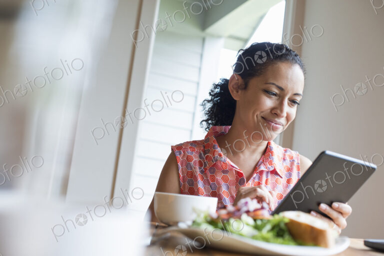 A young woman reading from the screen of a digital tablet, seated at a table. Coffee…