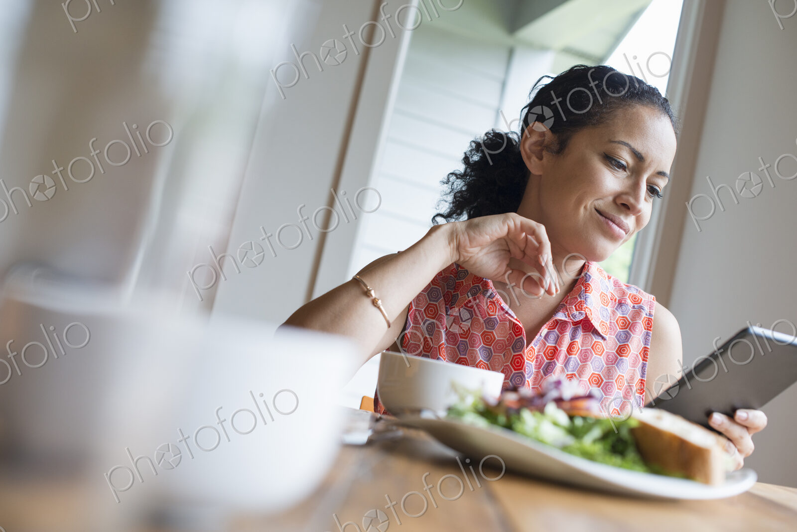 A young woman reading from the screen of a digital tablet, seated at a table. Coffee…