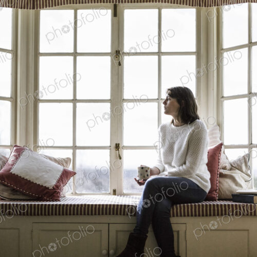 A young woman seated at a window seat, with a cup of tea.
