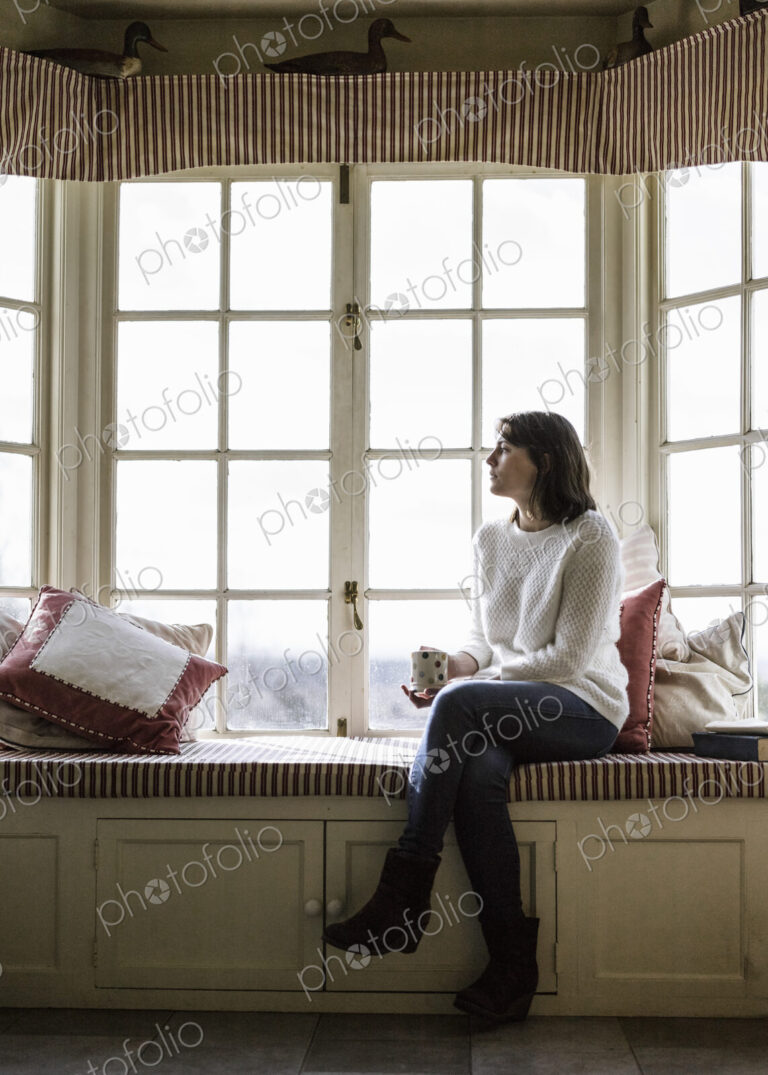 A young woman seated at a window seat, with a cup of tea.