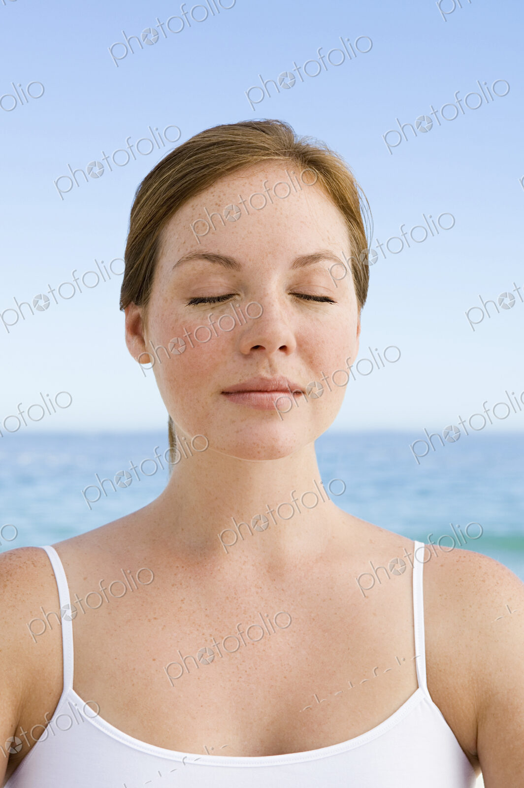A young woman seated on the beach in a relaxed pose, with her eyes closed.