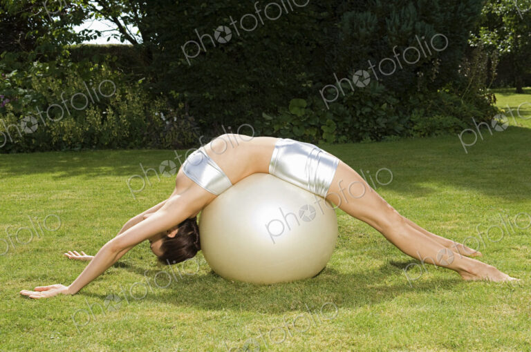 A young woman showing her flexibility, leaning backwards over an exercise ball, with…