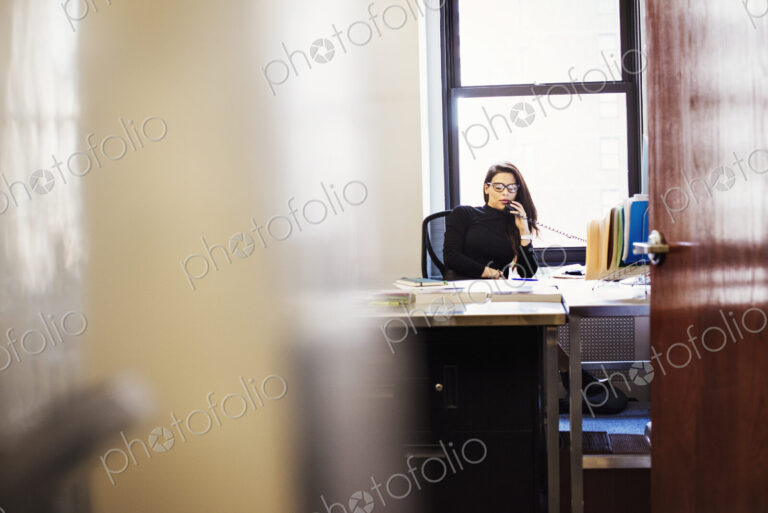 A young woman sitting at a desk in an office holding a telephone to her head.
