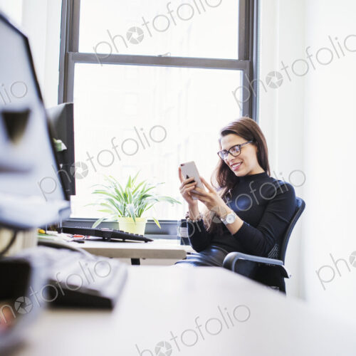 A young woman sitting at a desk in an office looking down at a cellphone. A young woman sitting at a desk in an office looking down at a cellphone.