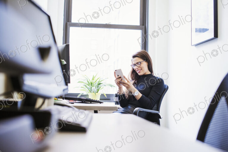A young woman sitting at a desk in an office looking down at a cellphone.