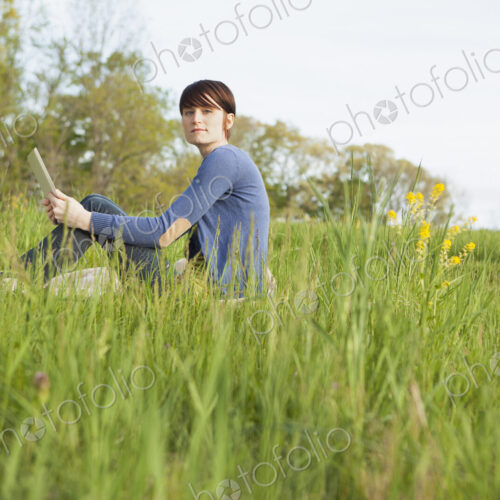 A young woman sitting in an open space, a grass field, on a blanket, holding a digital… A young woman sitting in an open space, a grass field, on a blanket, holding a digital…