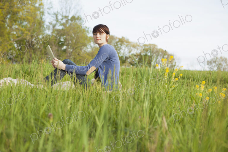 A young woman sitting in an open space, a grass field, on a blanket, holding a digital…