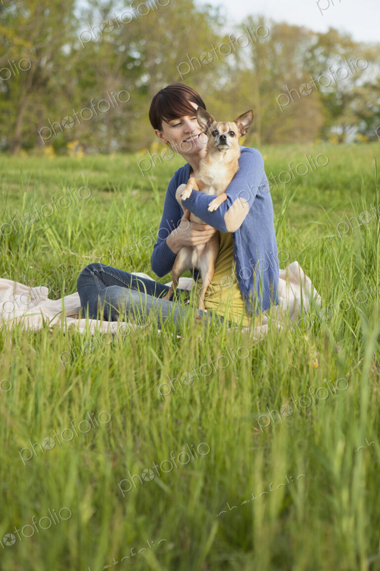 A young woman sitting in an open space, a grass field, on a blanket, holding a small…