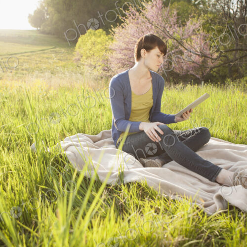 A young woman sitting in an open space, a grass field, on a blanket, holding and… A young woman sitting in an open space, a grass field, on a blanket, holding and…
