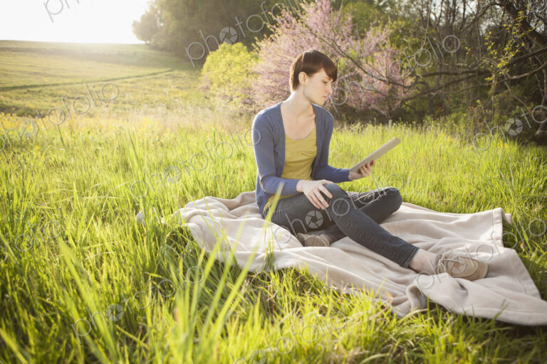 A young woman sitting in an open space, a grass field, on a blanket, holding and…