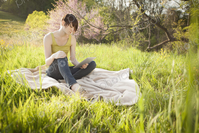 A young woman sitting in an open space, a grass field, on a blanket, reading from a…