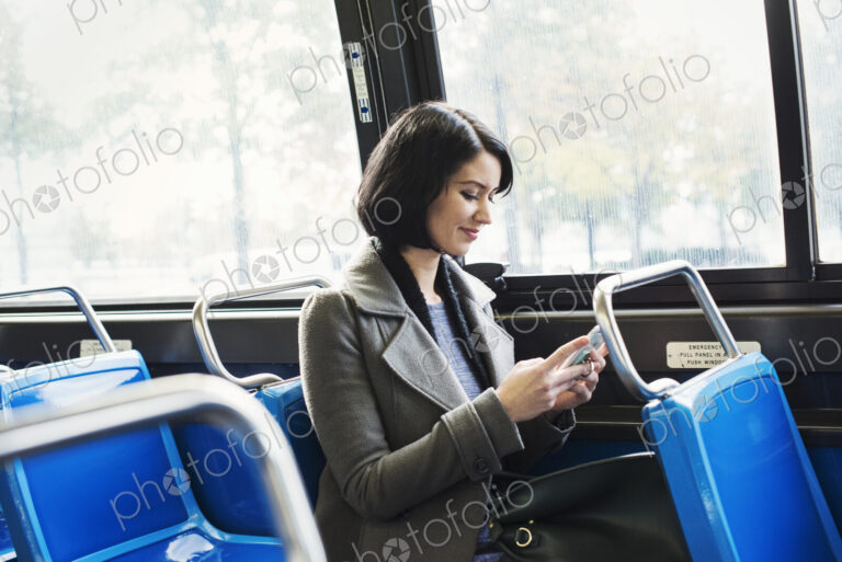 A young woman sitting on a train and looking down at a cellphone, smiling.