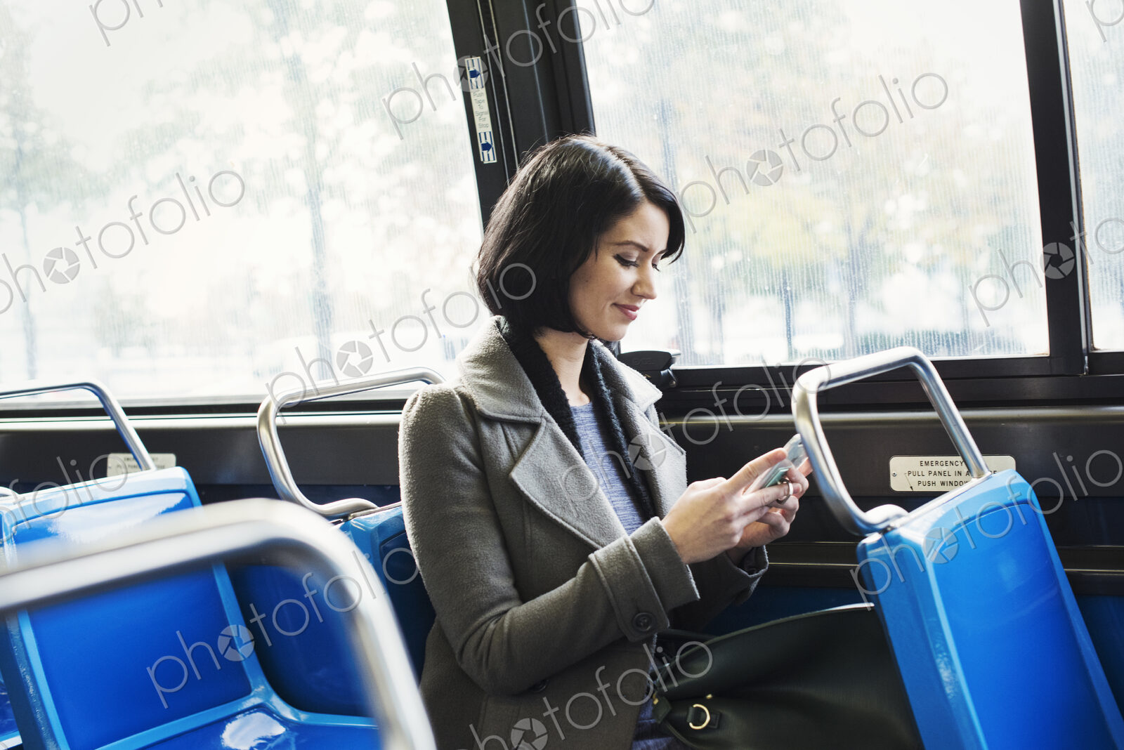 A young woman sitting on a train and looking down at a cellphone, smiling.