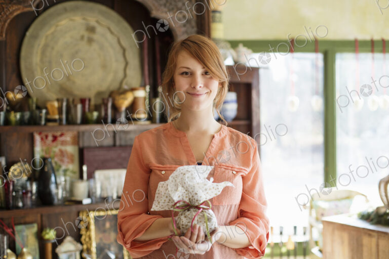 A young woman standing in a store full of antique objects, holding a tied packet with…