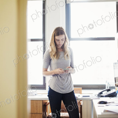 A young woman standing in an office looking down at her cellphone.