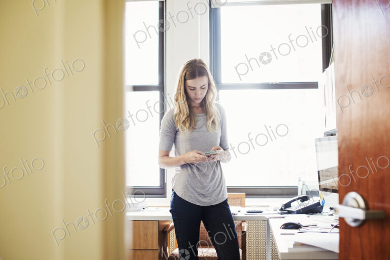 A young woman standing in an office looking down at her cellphone.