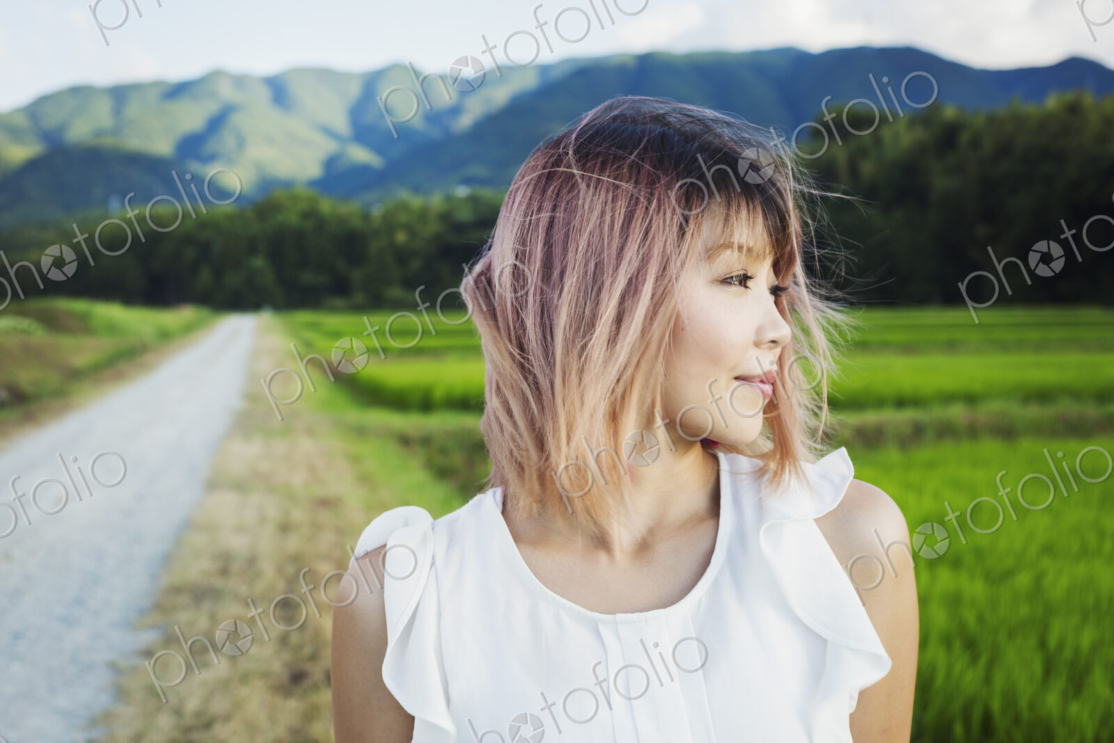 A young woman standing in open space by rice paddy fields of green shoots, and…