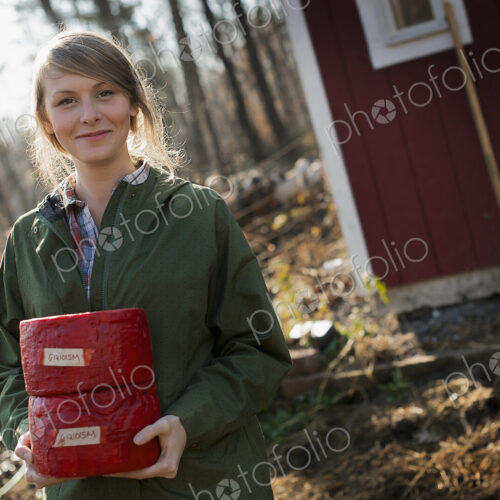 A young woman standing outside a dairy farm building holding two blocks of cheese.