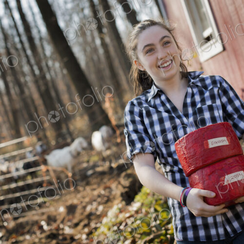 A young woman standing outside a dairy farm building holding two blocks of cheese.