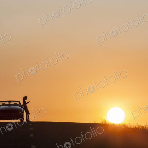 A young woman using her phone standing next to her car parked at sunset.