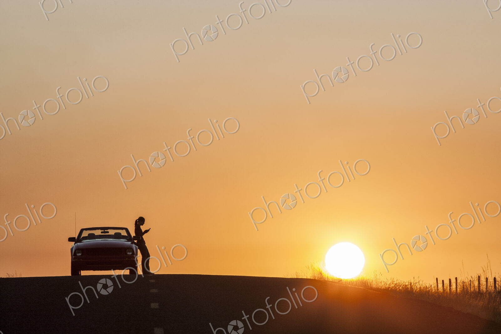 A young woman using her phone standing next to her car parked at sunset.