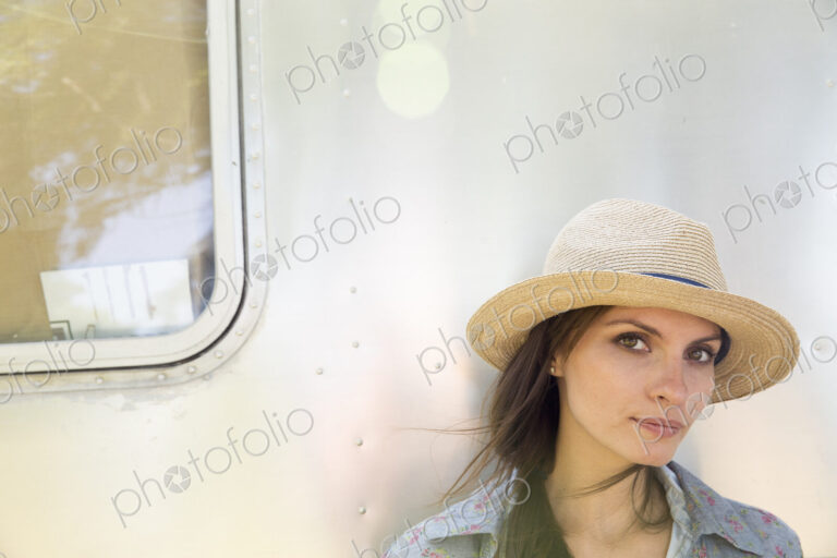 A young woman wearing a hat sitting in the shade of a silver coloured trailer.