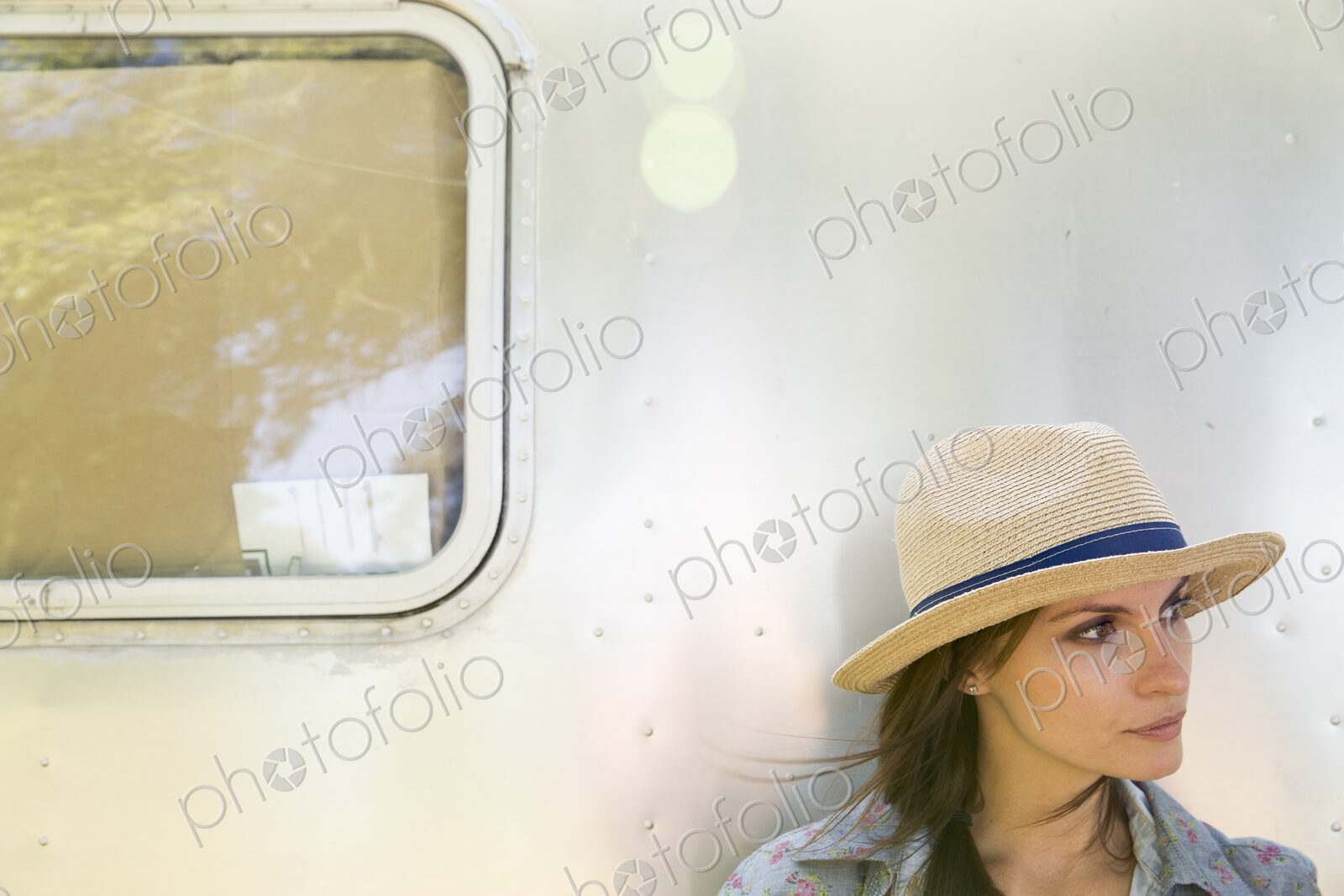 A young woman wearing a hat sitting in the shade of a silver trailer.