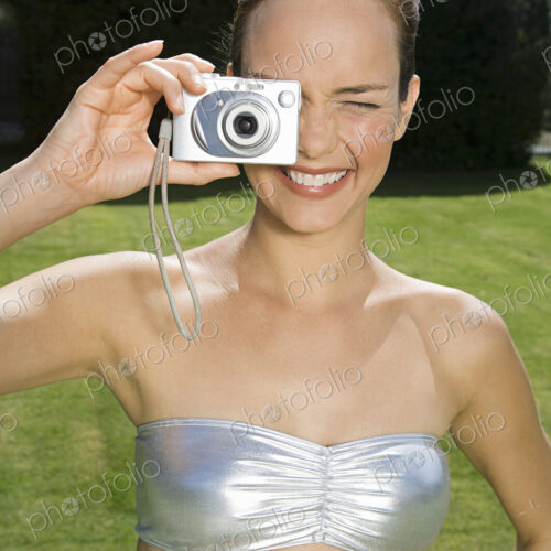 A young woman wearing a silver bandeau top, taking a photograph with a small camera.