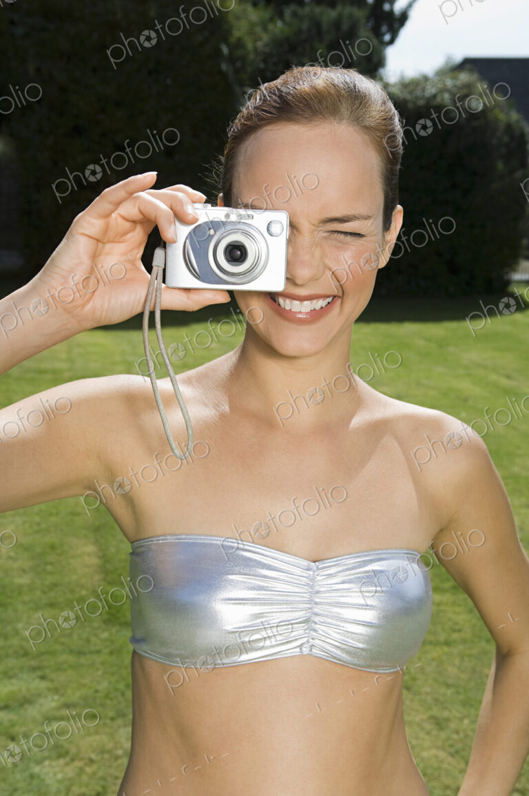 A young woman wearing a silver bandeau top, taking a photograph with a small camera.