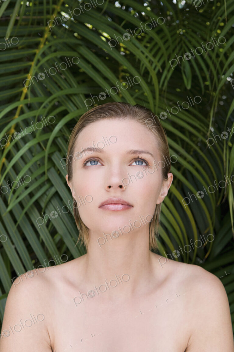A young woman with bare shoulders, looking upwards. Tropical plants in the background.