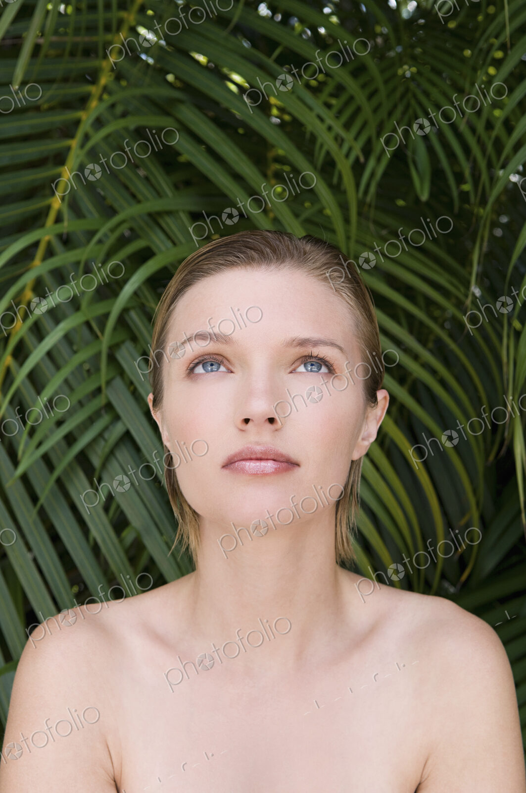 A young woman with bare shoulders, looking upwards. Tropical plants in the background.