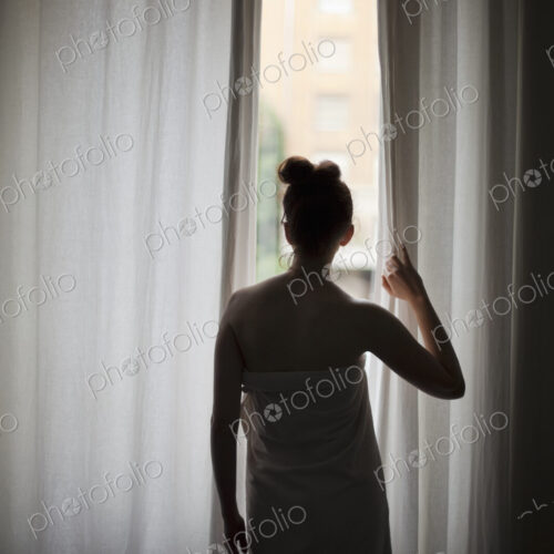A young woman with her hair up, wearing a bath towel, looking through long curtains at…