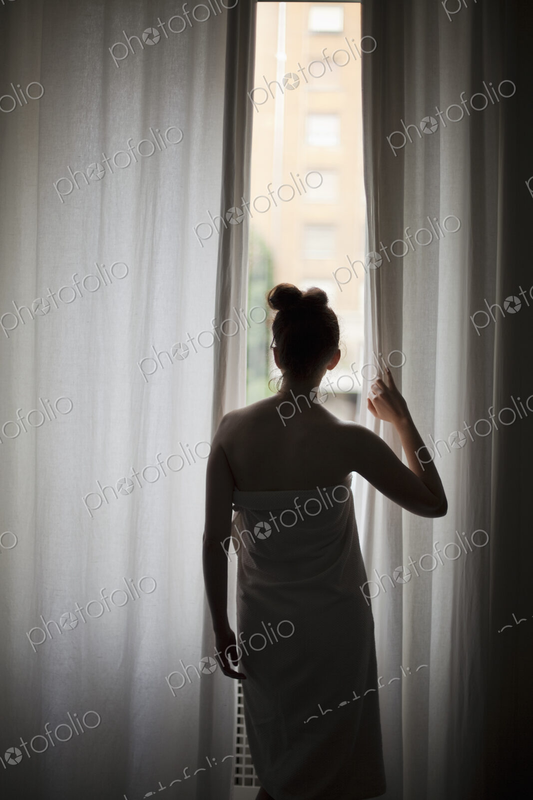 A young woman with her hair up, wearing a bath towel, looking through long curtains at…