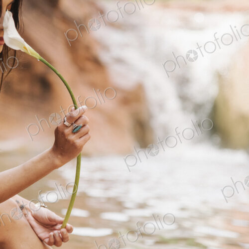 A young woman with long brown hair sitting by a river, smelling an Arum Lily. A young woman with long brown hair sitting by a river, smelling an Arum Lily.