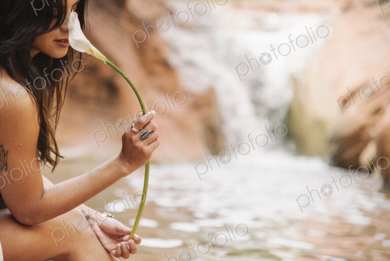 A young woman with long brown hair sitting by a river, smelling an Arum Lily.