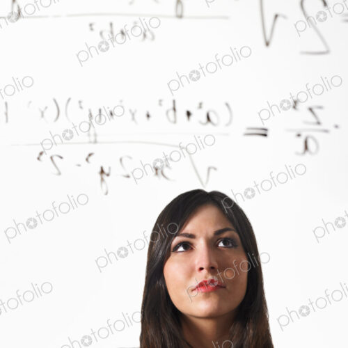 A young woman writing a mathematical equation with black marker on a clear seethrough…
