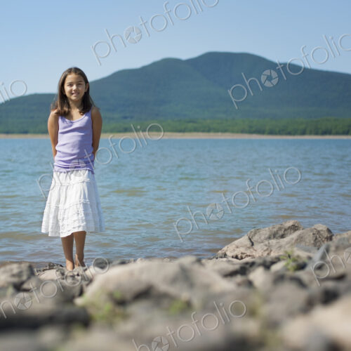 A youngirl in a white skirt and purple top. Standing on the shore of a lake.