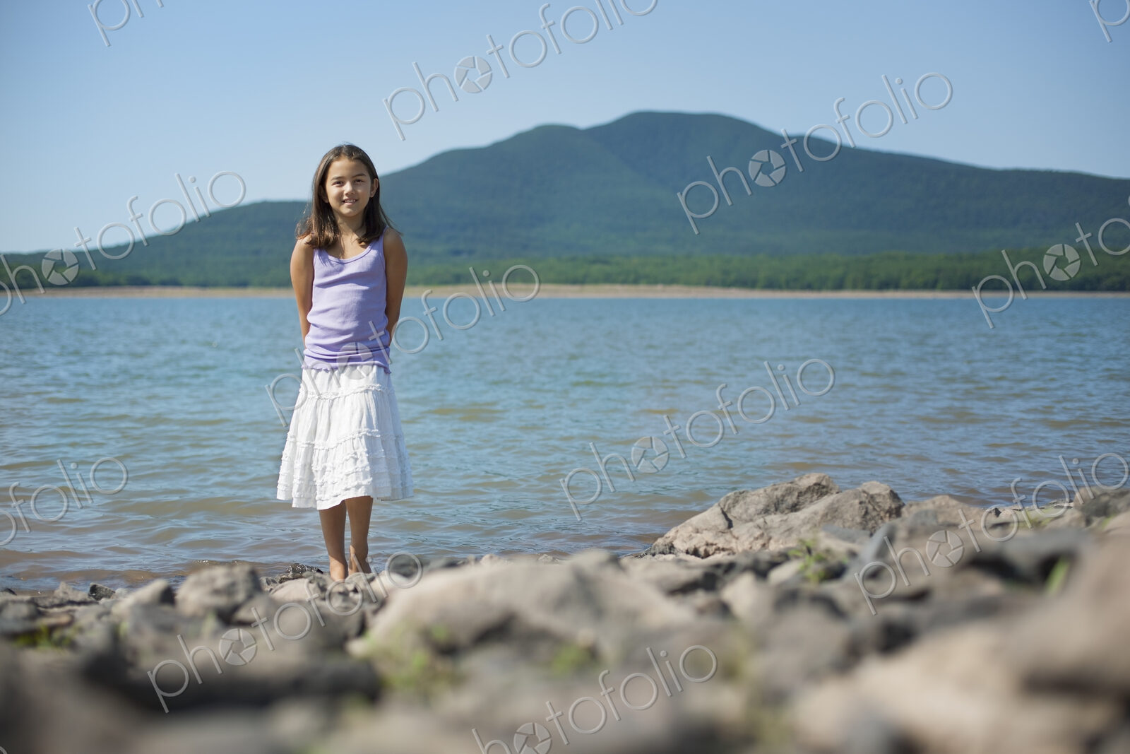 A youngirl in a white skirt and purple top. Standing on the shore of a lake.
