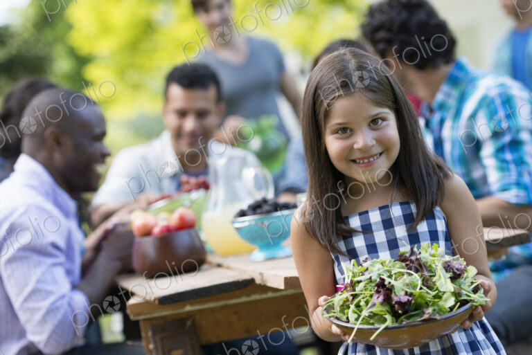 Adults and children around a table in a garden. A child holding a bowl of salad.